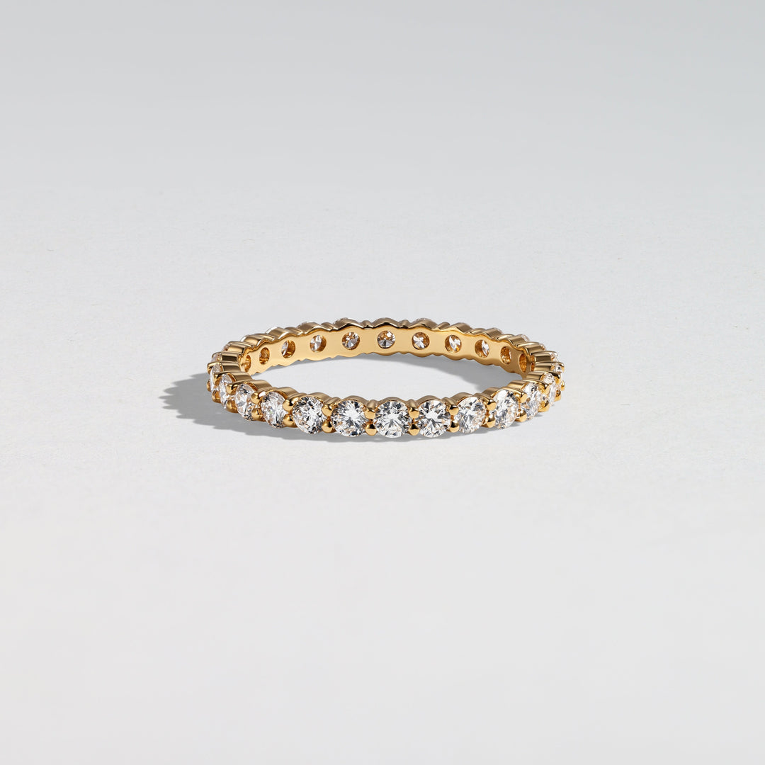 A gold eternity ring set with round-cut clear gemstones, photographed on a clean light gray background #material_gold
