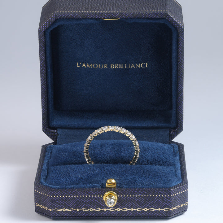 A diamond-encrusted ring displayed inside an open dark blue jewelry box with gold trim and the text 'L'AMOUR BRILLIANCE' on the inner lid, set against a light gray studio background #material_gold