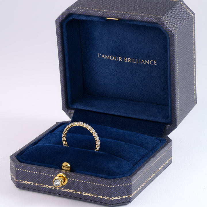 A gold ring with a pavé diamond band displayed inside an open dark blue textured jewelry box with gold trim and the brand name 'L'AMOUR BRILLIANCE' printed on the inner lid, set against a clean white background. #material_gold