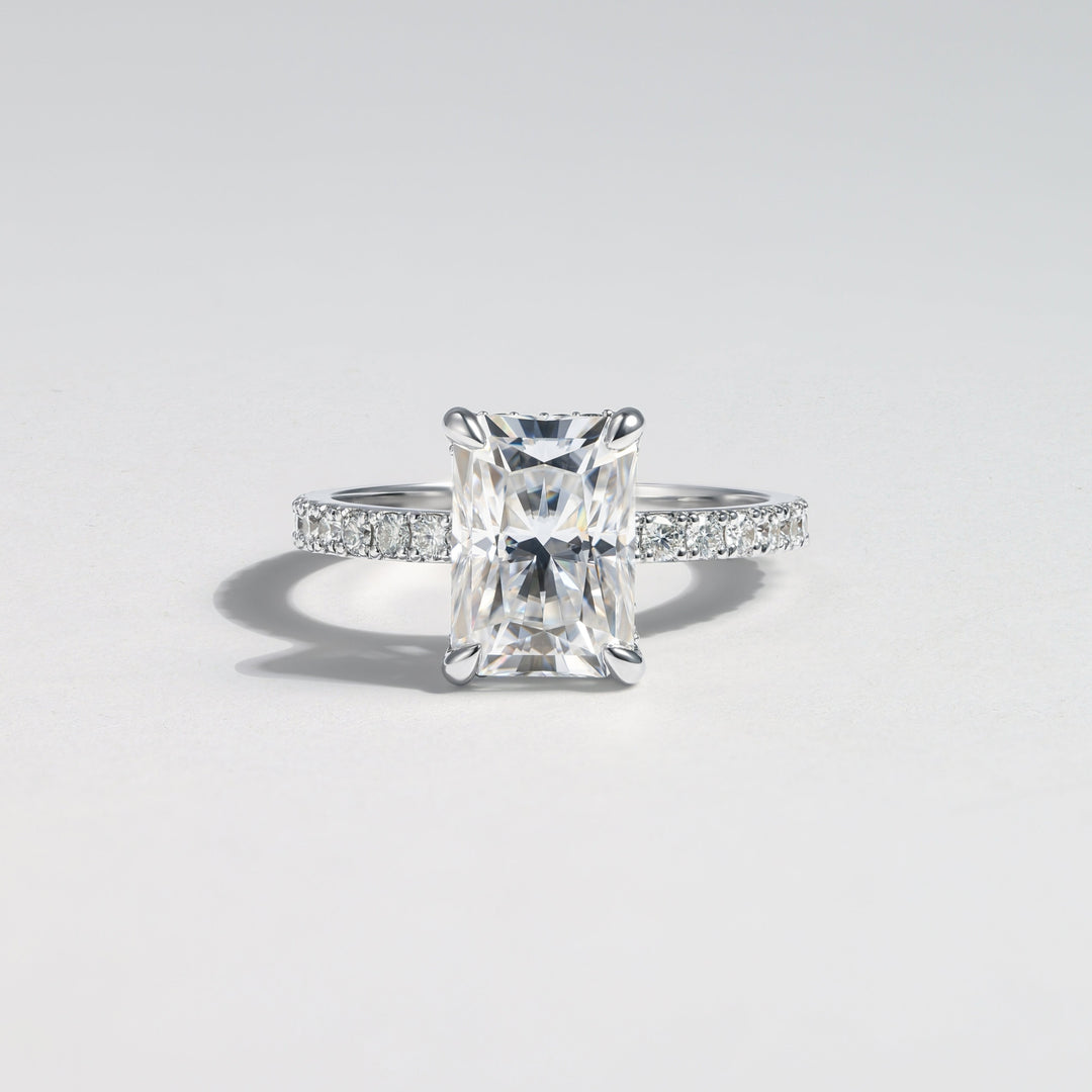 A radiant cut diamond engagement ring with a silver-colored band accented by small pavé diamonds, centered on a light gray background #material_white