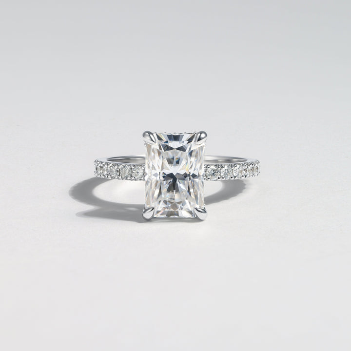 A radiant cut diamond engagement ring with a silver-colored band accented by small pavé diamonds, centered on a light gray background #material_white