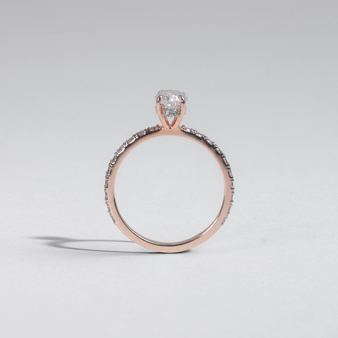 A rose gold ring with a single round-cut clear gemstone and a pavé band, photographed on a light gray background #material_rose