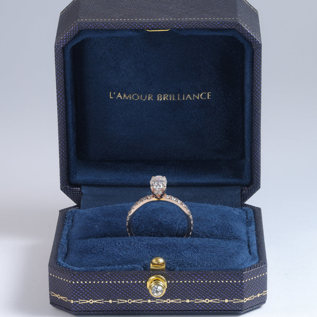 A diamond ring with a gold band displayed inside an open dark blue jewelry box with gold trim and the text 'L'AMOUR BRILLIANCE' on the inner lid, set against a light gray studio background #material_rose