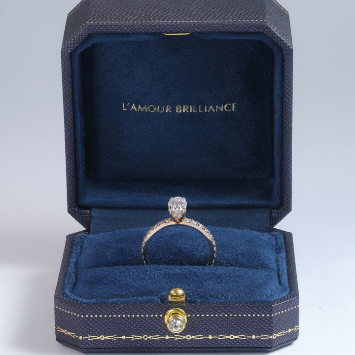 A diamond ring with a gold band displayed inside an open dark blue jewelry box with gold trim and the text 'L'AMOUR BRILLIANCE' on the inner lid, set against a light gray studio background #material_rose