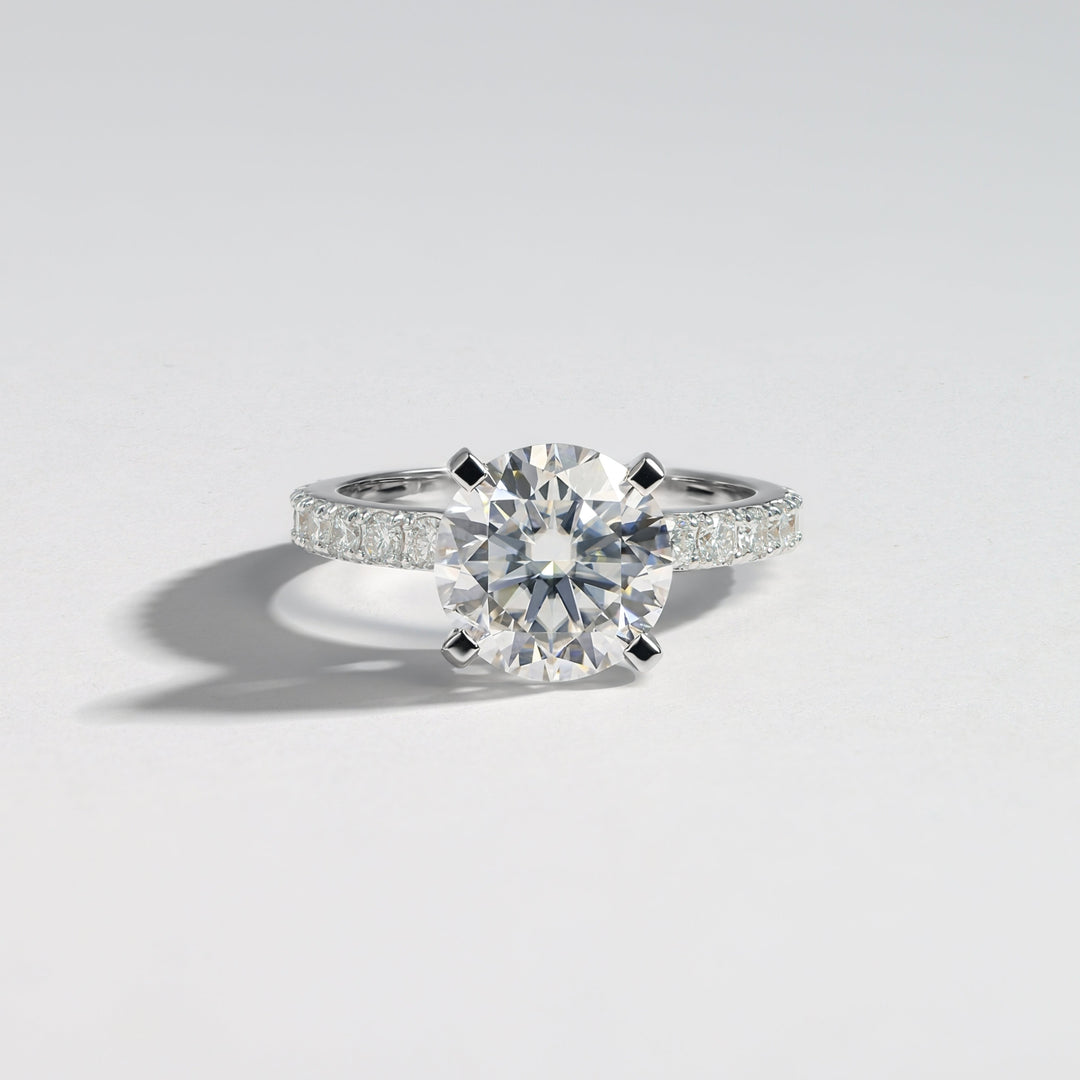 A classic solitaire engagement ring featuring a round brilliant-cut center stone in a four-prong setting on a silver-colored band adorned with pavé side stones, photographed on a clean light gray background #material_white