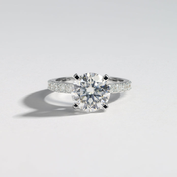 A classic solitaire engagement ring featuring a round brilliant-cut center stone in a four-prong setting on a silver-colored band adorned with pavé side stones, photographed on a clean light gray background #material_white