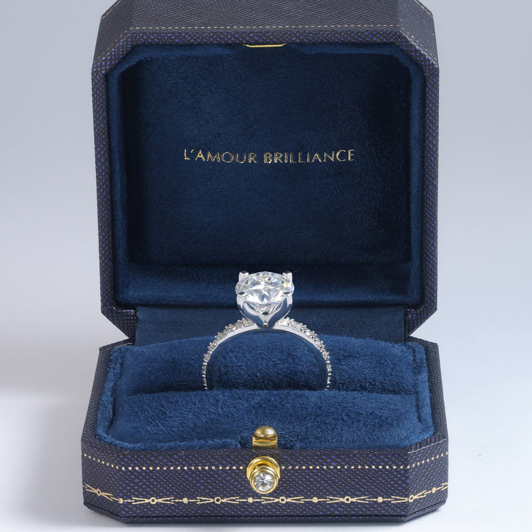 A silver engagement ring with a round-cut center stone and small side stones, displayed inside an open dark blue velvet jewelry box with gold trim. The inside lid is lined with dark blue velvet and features the gold text 'L'AMOUR BRILLIANCE' #material_white