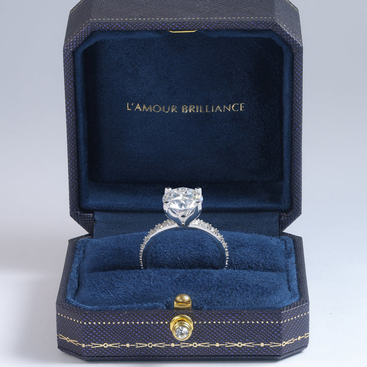 A silver engagement ring with a round-cut center stone and small side stones, displayed inside an open dark blue velvet jewelry box with gold trim. The inside lid is lined with dark blue velvet and features the gold text 'L'AMOUR BRILLIANCE' #material_white
