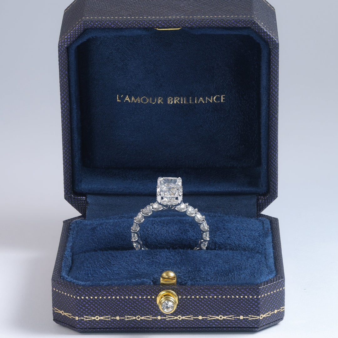 A diamond ring displayed in an open dark blue jewelry box with gold trim and the text 'L'AMOUR BRILLIANCE' on the inside lid, photographed on a light gray studio background #material_white