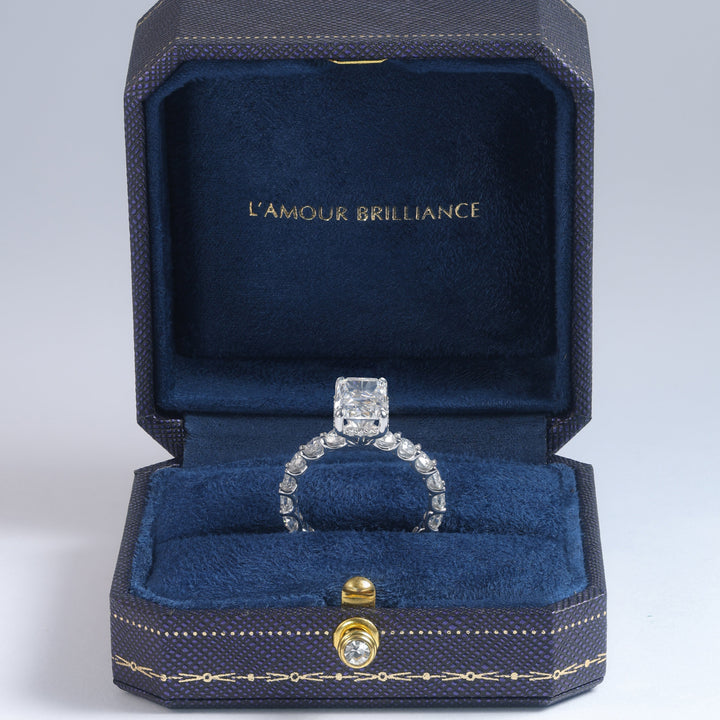 A diamond ring displayed in an open dark blue jewelry box with gold trim and the text 'L'AMOUR BRILLIANCE' on the inside lid, photographed on a light gray studio background #material_white