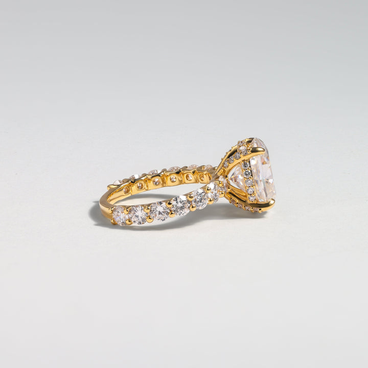 A yellow gold ring featuring a large cushion-cut clear center stone and a pavé-set band with small clear stones, photographed on a light gray gradient background #material_gold