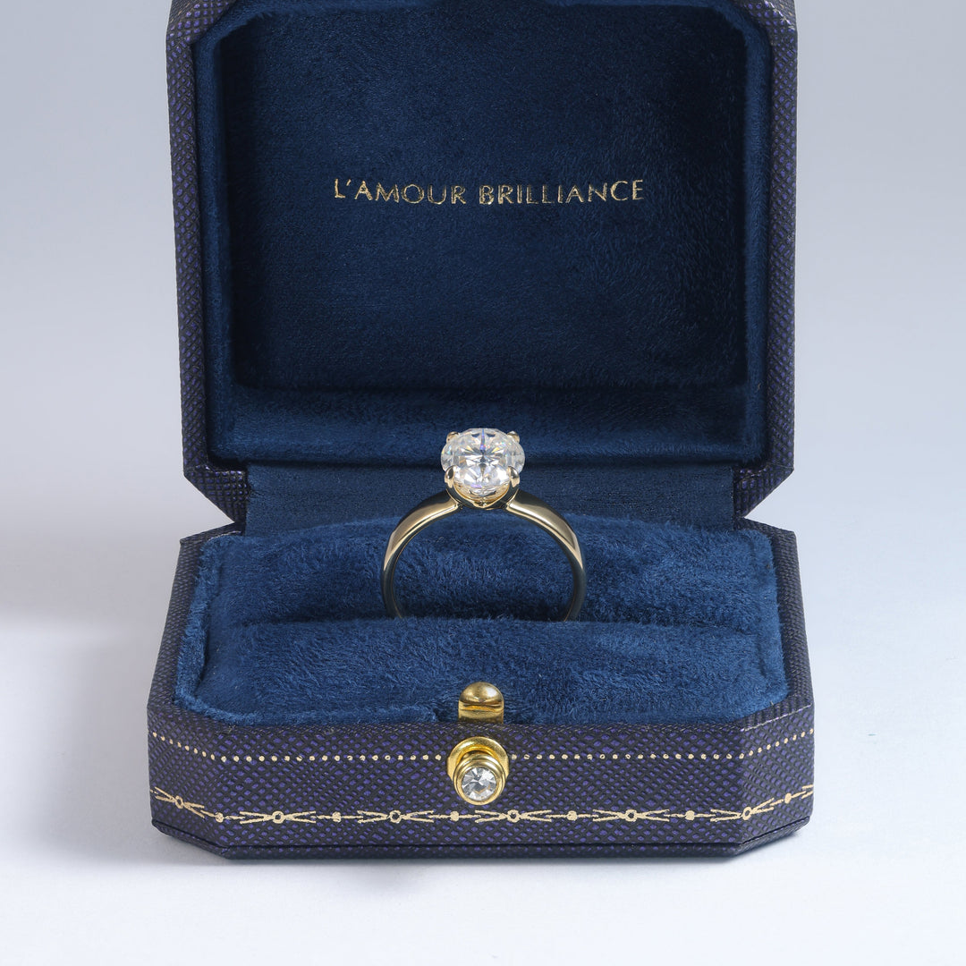 A yellow gold ring with a round-cut clear gemstone displayed inside an open dark blue velvet jewelry box with gold trim. The inside lid features the gold-embossed brand name 'L'AMOUR BRILLIANCE #material_gold