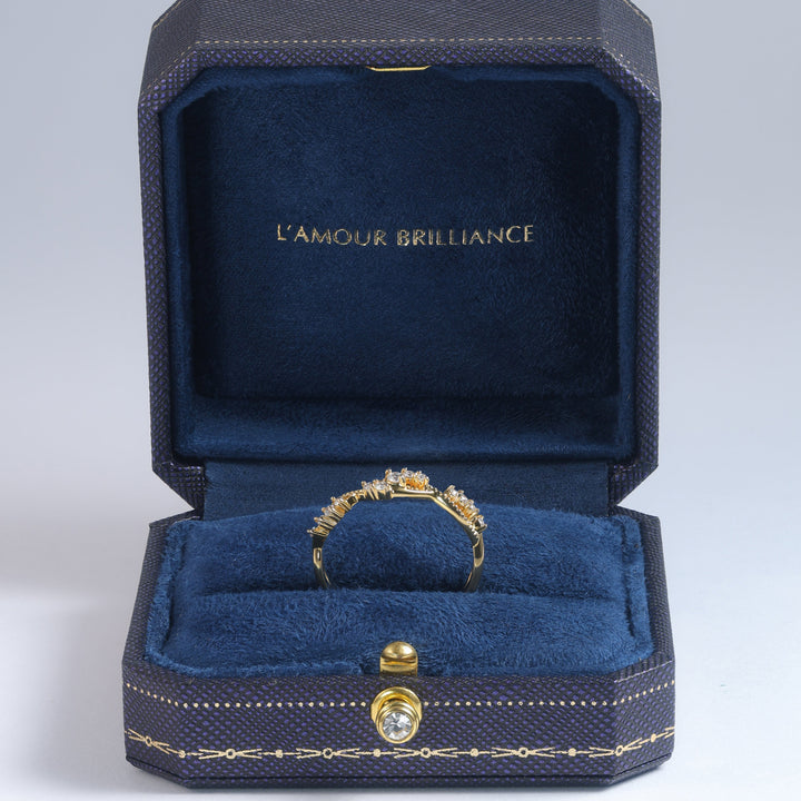 A yellow gold ring with a round-cut clear gemstone displayed inside an open dark blue velvet jewelry box with gold trim. The inside lid features the gold-embossed brand name 'L'AMOUR BRILLIANCE' #material_gold