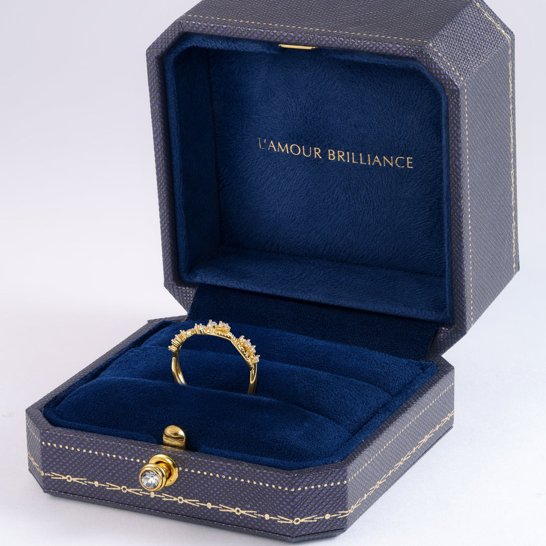 An open dark textured jewelry box with a deep blue velvet interior, displaying a gold ring with a small clear gemstone. The inside lid is lined with matching blue velvet and features the gold text 'L'AMOUR BRILLIANCE' #material_gold
