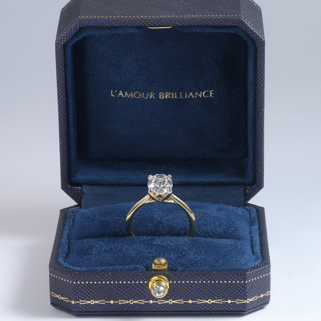 A yellow gold solitaire ring with a round brilliant-cut diamond is displayed inside an open dark blue velvet jewelry box with gold trim. The inside lid features the gold-foiled text 'L'AMOUR BRILLIANCE' #material_gold