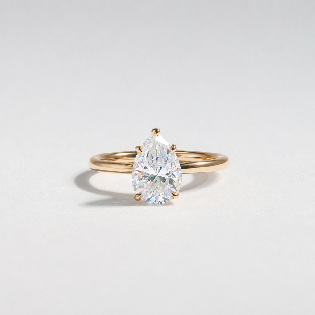 A pear-cut diamond engagement ring in a yellow gold band, photographed on a clean, light gray studio background. #material_gold