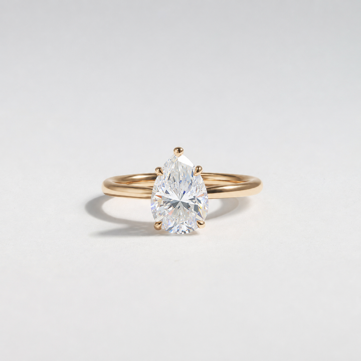 A pear-cut diamond engagement ring in a yellow gold band, photographed on a clean, light gray studio background. #material_gold