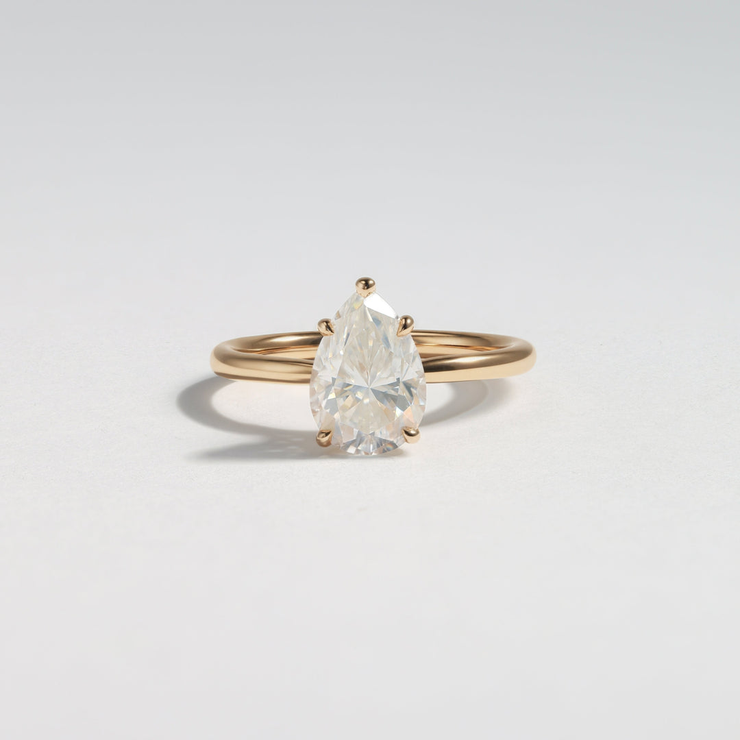 A pear-cut clear gemstone ring set on a slim yellow gold band, photographed on a clean light gray background #material_gold