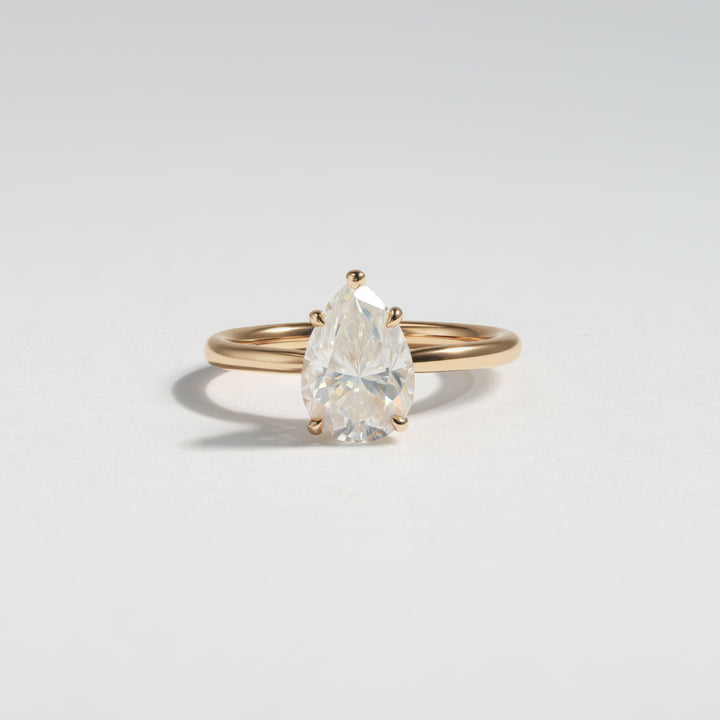 A pear-cut clear gemstone ring set on a slim yellow gold band, photographed on a clean light gray background #material_gold