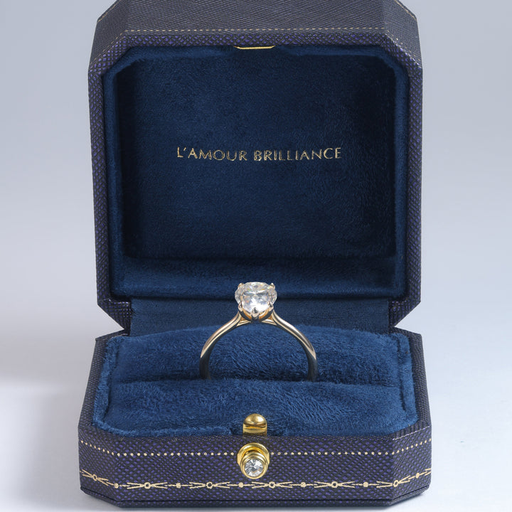 A yellow gold ring with a round clear gemstone displayed inside an open dark blue velvet jewelry box. The inside lid features the gold-foiled text 'L'AMOUR BRILLIANCE' #material_gold