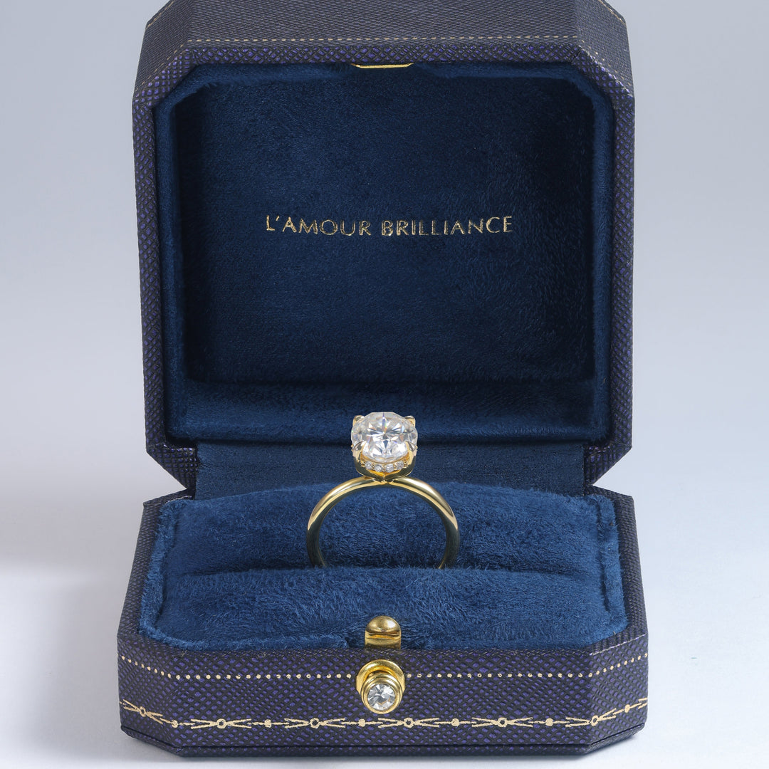 A yellow gold ring with a round clear gemstone displayed inside an open dark blue velvet jewelry box. The inside lid features the gold-foiled text 'L'AMOUR BRILLIANCE' #material_gold