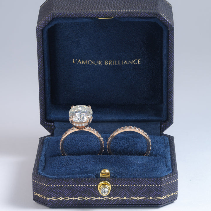 An open dark blue jewelry box with gold trim and the words 'L'AMOUR BRILLIANCE' printed inside, holding two rings. One ring has a large clear gemstone in a silver-toned setting, and the other has a pinkish gemstone in a rose-gold-toned setting #material_rose