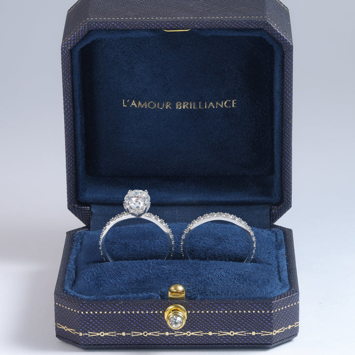 An open dark blue jewelry box with gold trim and the words 'L'AMOUR BRILLIANCE' printed inside, holding two rings on a blue velvet cushion against a light gray studio background #material_white