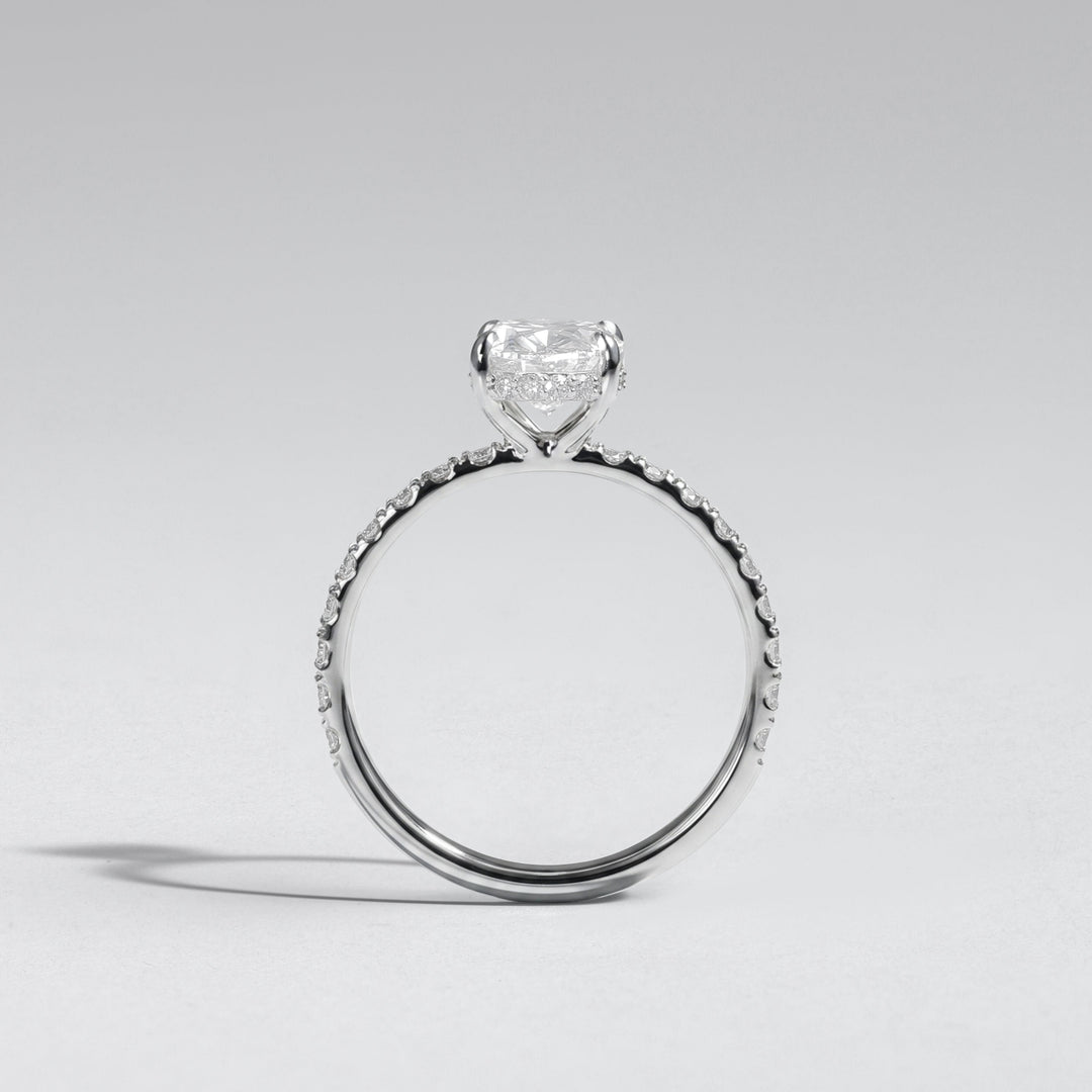 A silver-toned solitaire engagement ring featuring a heart-shaped clear gemstone in a prong setting, with a textured pavé band, shown on a neutral light gray background #material_white