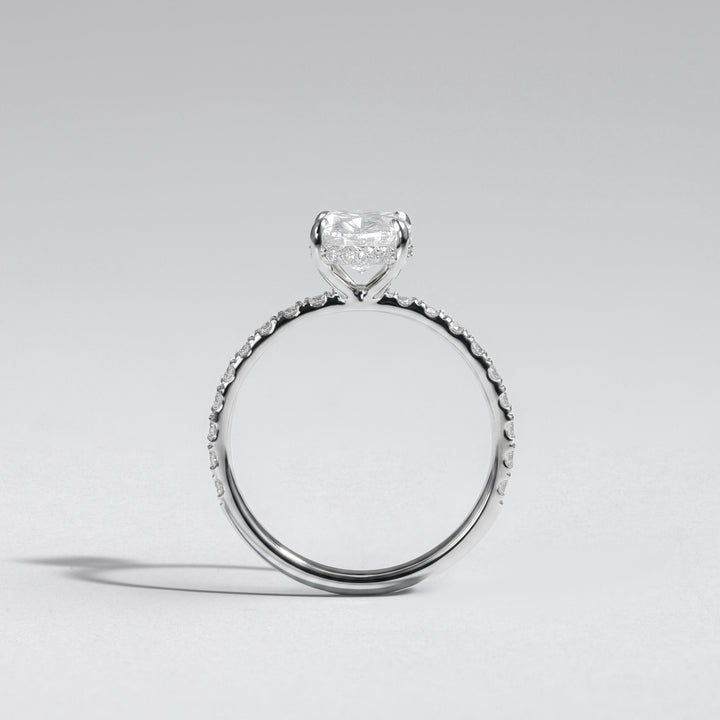 A silver-toned solitaire engagement ring featuring a heart-shaped clear gemstone in a prong setting, with a textured pavé band, shown on a neutral light gray background #material_white