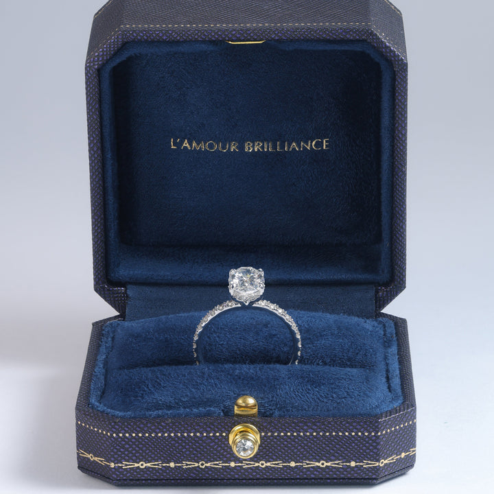 A diamond engagement ring with a silver-toned band displayed inside an open dark blue velvet jewelry box. The inside lid features the gold-foiled text 'L'AMOUR BRILLIANCE' #material_white