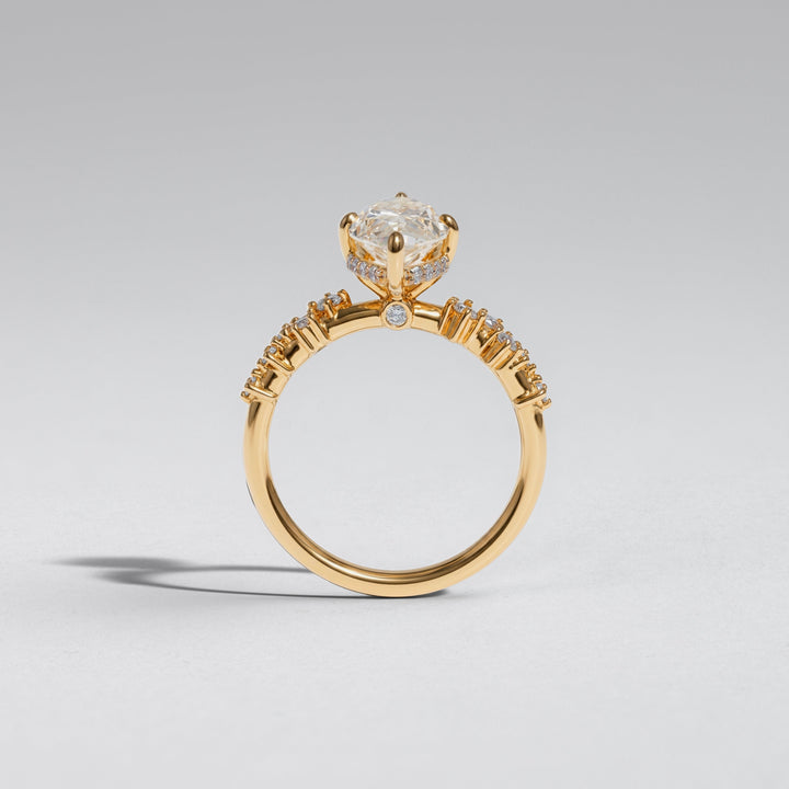 A yellow gold ring featuring a large clear round-cut center stone in a prong setting, with small clear stones along the shoulders, shown on a neutral light gray background #material_gold