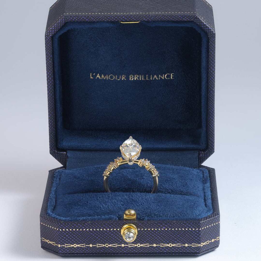 A yellow gold ring with a large clear marquise-cut diamond is displayed inside an open dark blue velvet jewelry box with gold trim. The inside lid of the box features the gold-embossed brand name 'L'AMOUR BRILLIANCE' #material_gold