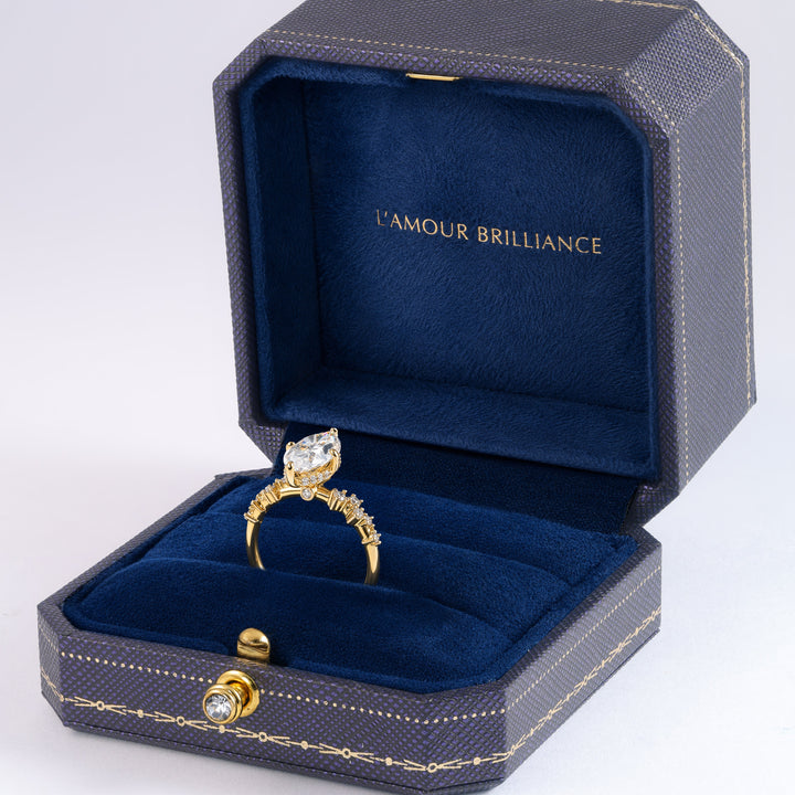 An open dark textured jewelry box with deep blue velvet lining, featuring the gold-foiled brand name 'L'AMOUR BRILLIANCE' on the inner lid. A yellow gold ring with a large clear gemstone and smaller side stones is displayed inside the box #material_gold