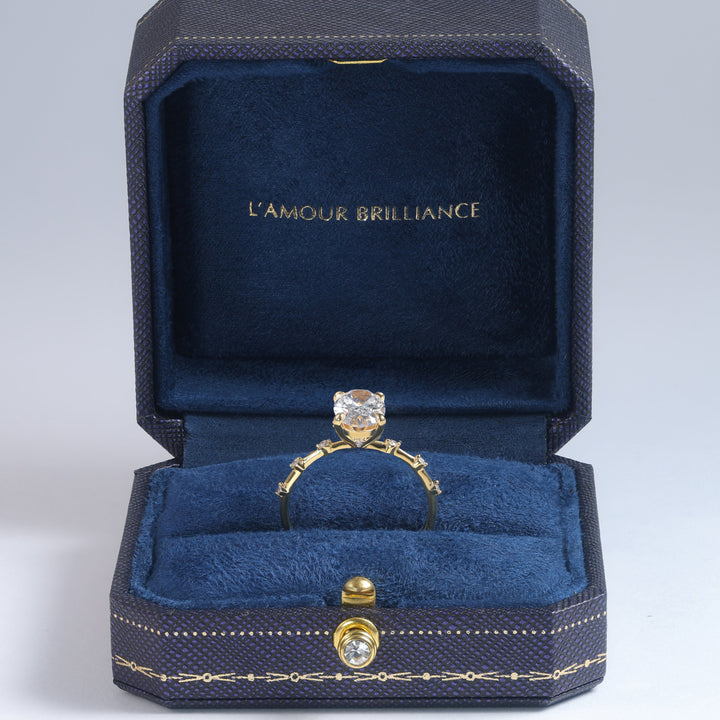 A yellow gold ring with a round-cut clear gemstone displayed inside an open dark blue velvet jewelry box. The inside lid features the gold-foiled text 'L'AMOUR BRILLIANCE' #material_gold