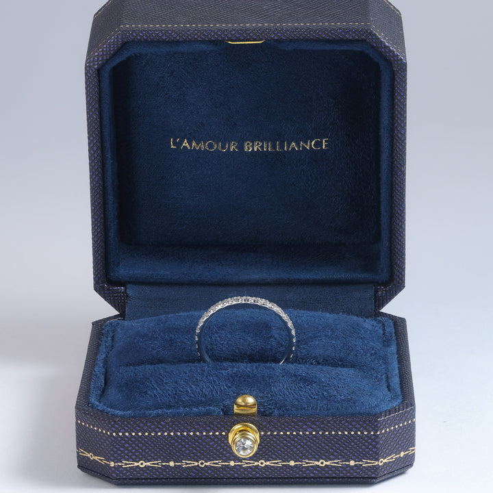 A silver band ring with a pavé diamond setting, displayed inside an open luxury jewelry box. The box features a deep blue velvet interior with gold lettering that reads 'L'AMOUR BRILLIANCE' on the inner lid, and a small gold clasp on the front #material_white