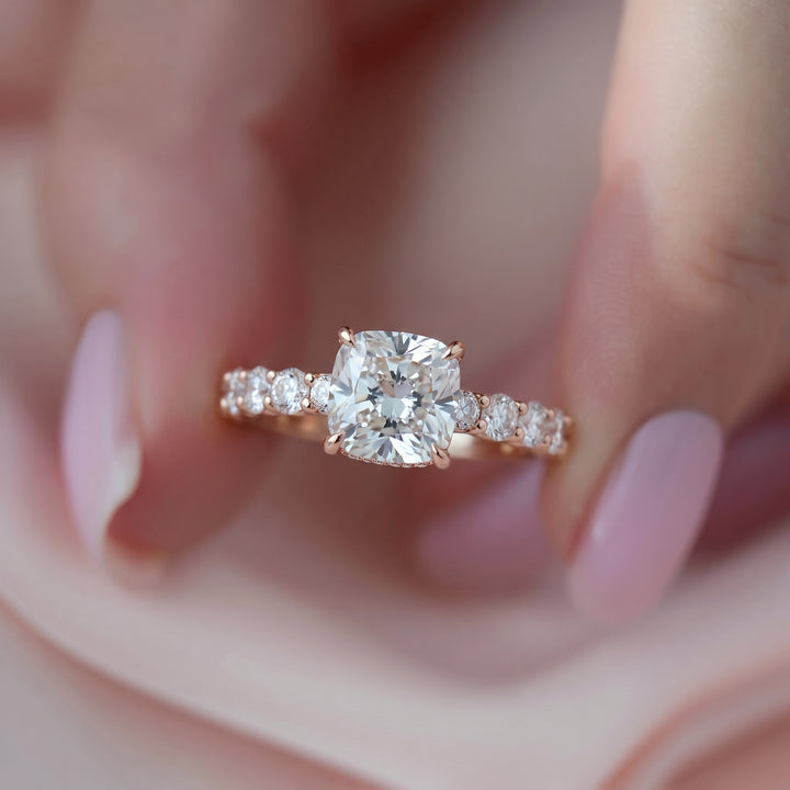 A close-up of a rose gold engagement ring with a cushion-cut center stone and smaller side stones, held on a finger with manicured pink nails. #material_rose