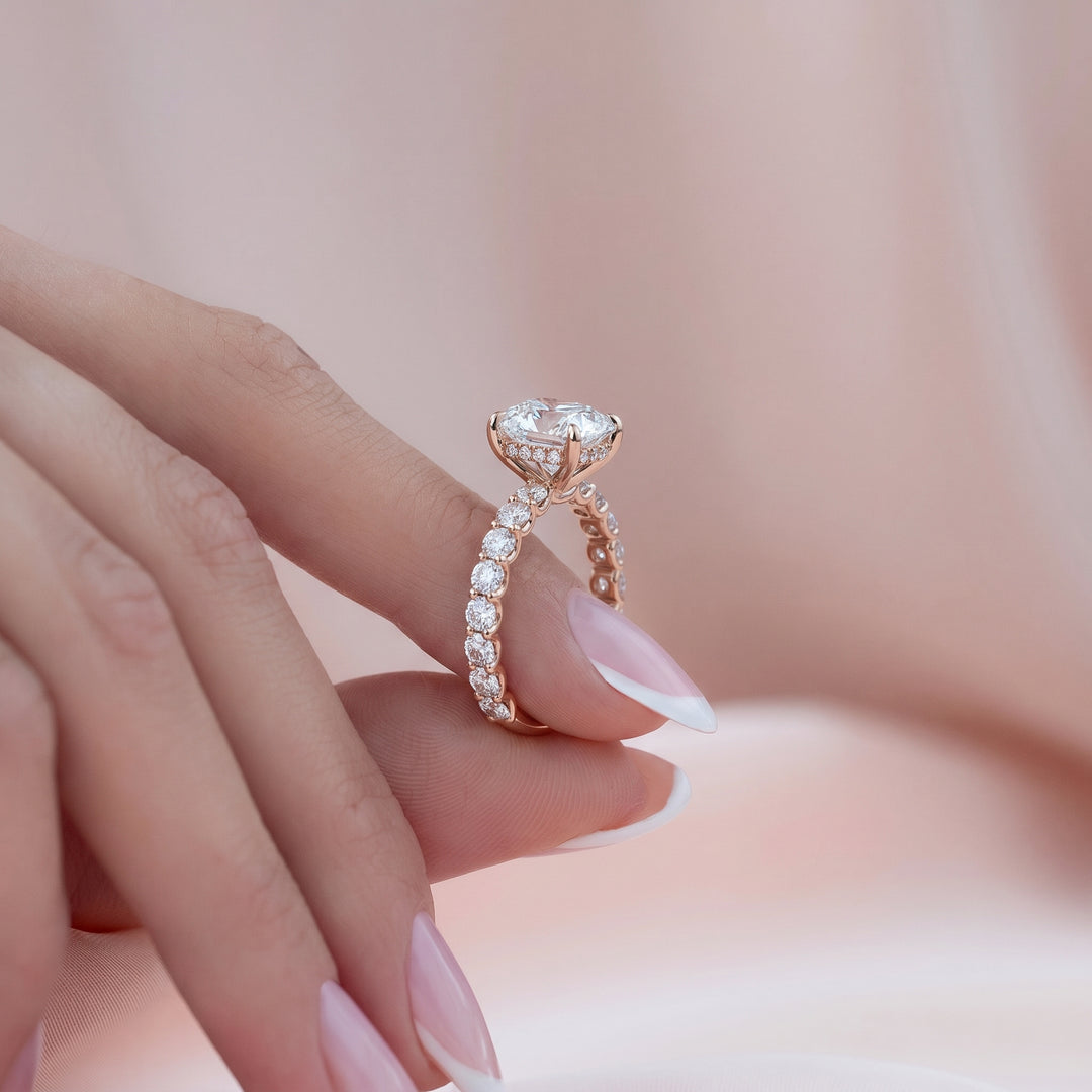 A close-up lifestyle shot of a rose gold engagement ring with a round-cut diamond and a pavé-set band of small diamonds, worn on a hand with French-tipped nails against a soft pink background. #material_rose