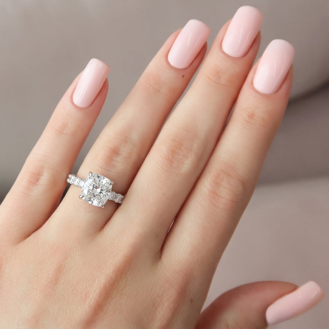 A close-up of a hand with pale pink manicured nails wearing a silver-toned ring with a large clear cushion-cut center stone and small side stones, set against a soft neutral background. #material_white