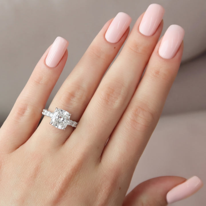 A close-up of a hand with pale pink manicured nails wearing a silver-toned ring with a large clear cushion-cut center stone and small side stones, set against a soft neutral background. #material_white