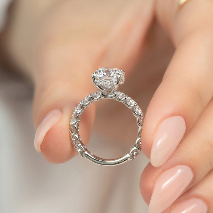 A silver-toned ring with a heart-shaped clear gemstone and small pavé-set stones along the band, held between a person's fingers.#material_white