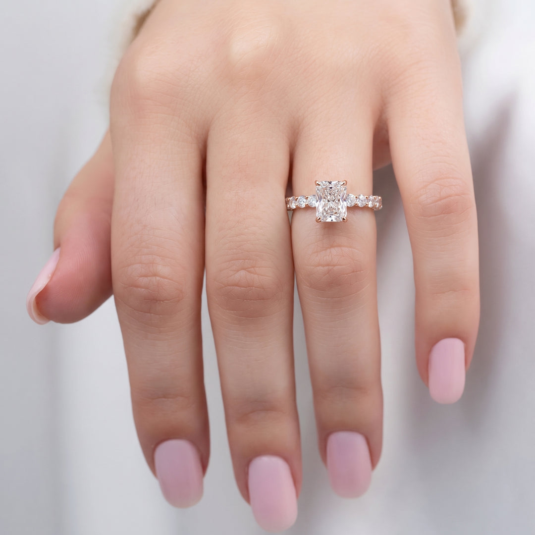 A close-up of a hand wearing a rose gold engagement ring with a large round-cut center stone and smaller side stones on a soft light gray background. #material_rose