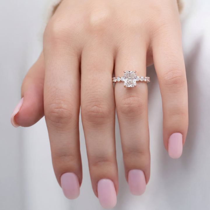 A close-up of a hand wearing a rose gold engagement ring with a large round-cut center stone and smaller side stones on a soft light gray background. #material_rose