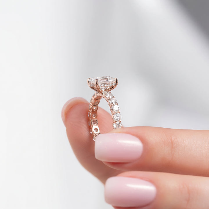A close-up of a rose gold engagement ring with a round-cut center stone and a pavé-set band, held between a person's fingers against a soft, light gray background. #material_rose