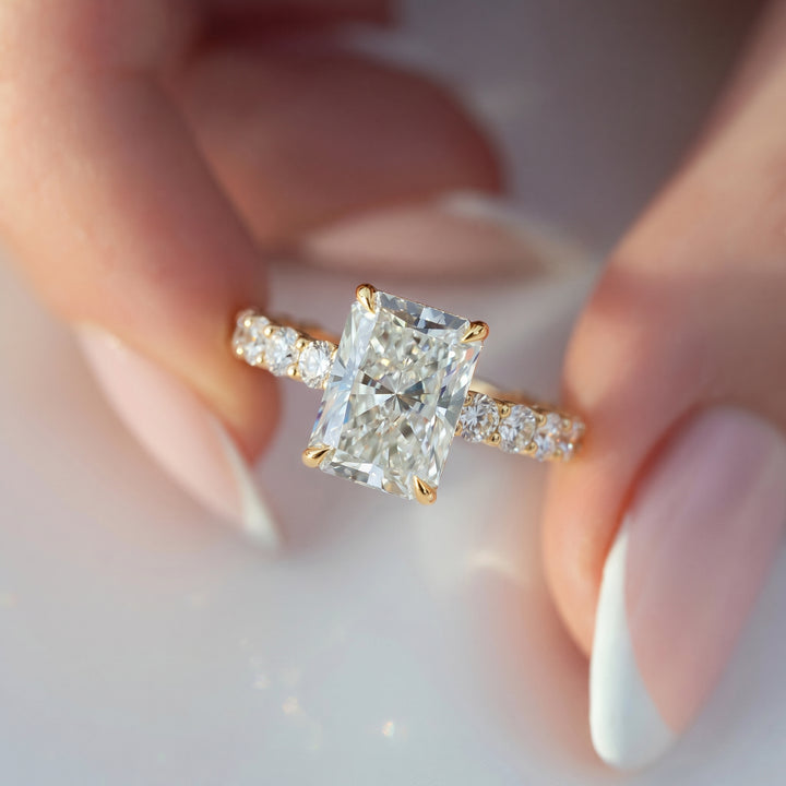 A close-up of a yellow gold engagement ring with a large rectangular brilliant-cut center stone and a pavé band, held between fingers with a soft-focus background. #material_gold