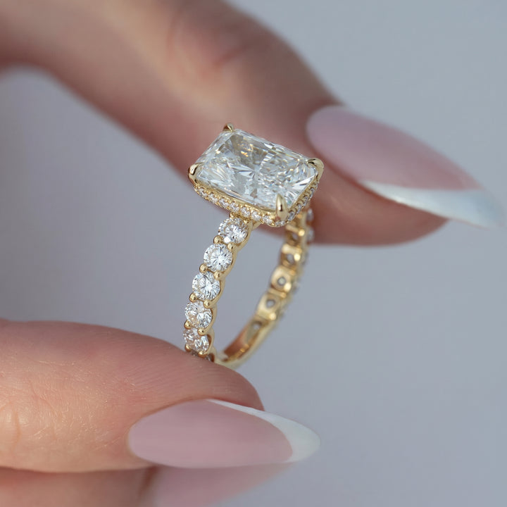 A close-up of a hand with a French manicure holding a yellow gold ring featuring a large rectangular clear gemstone and a pavé band with smaller round diamonds, set against a soft light gray background. #material_gold