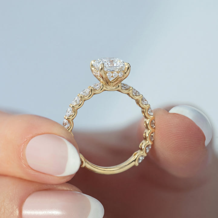 A close-up of a gold ring with a large clear center stone and small clear stones along the band, held between fingers against a soft light blue background. #material_gold