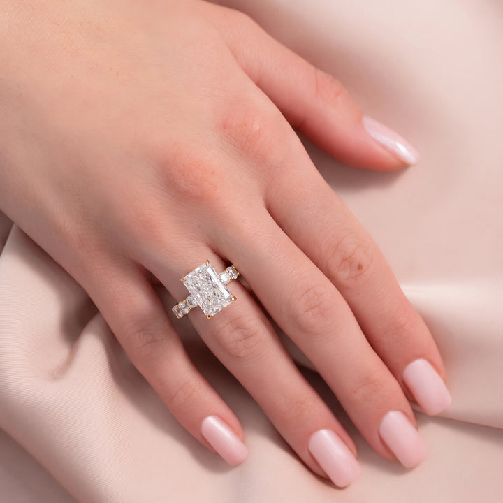 A close-up of a hand with pale pink manicured nails wearing a silver ring with a large rectangular clear gemstone, resting on a soft beige fabric background. #material_gold