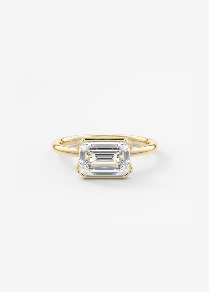 A yellow gold ring featuring an emerald-cut clear gemstone in a bezel setting, photographed against a clean, light gray background.#material_gold