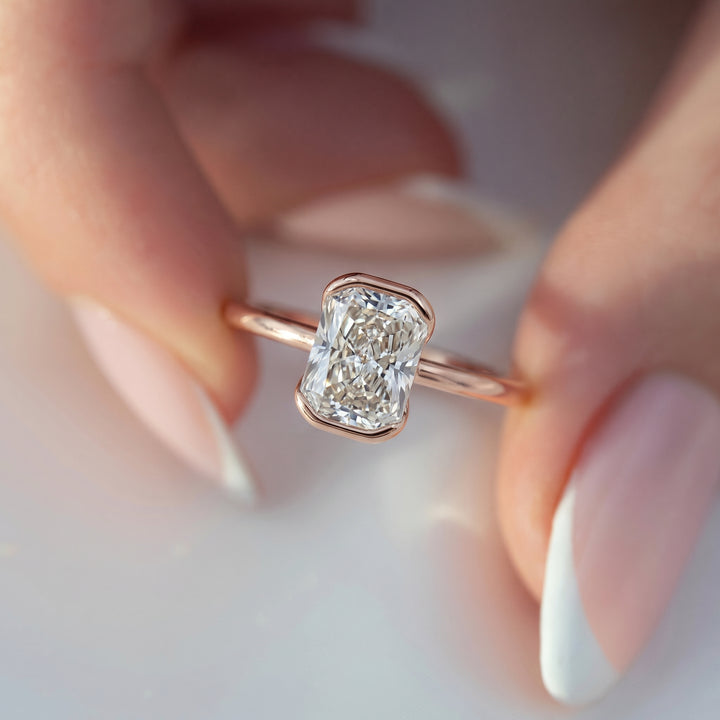 A close-up lifestyle shot of a rose gold ring with a rectangular emerald-cut diamond, held between a person's fingers with a soft-focus background. #material_rose