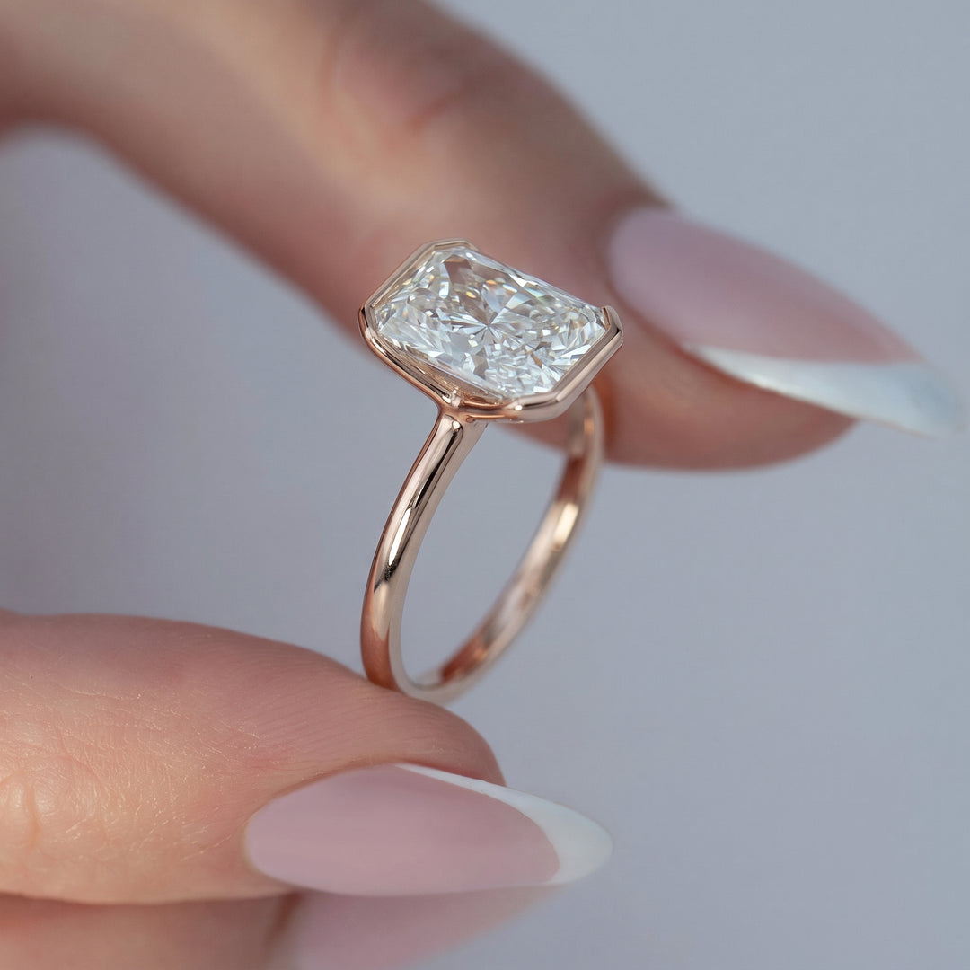 A close-up of a hand with French-tipped nails holding a rose gold ring with a large rectangular emerald-cut diamond. The diamond is clear and does not contribute to the color mapping according to the schema rules.#material_rose